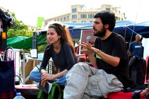 Enric Duran, seated, speaks into a microphone in his hand. Núria Güell, also seated, sits to his left. Tents in the background.