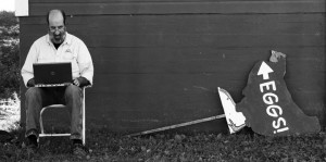 a blogger works at a laptop outside, to the left of a fallen over sign of a hen with the word Eggs! printed on her