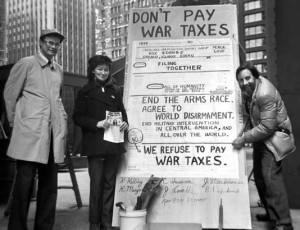 Brad Lyttle, Kathy Kelly, and Karl Meyer alongside a large sign reading 'Don’t Pay War Taxes; End the arms race; agree to world disarmament; end military intervention in central America and all over the world. We refuse to pay war taxes.'