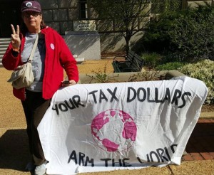 Carol Coney flashes peace sign next to banner reading “your tax dollars arm the world”