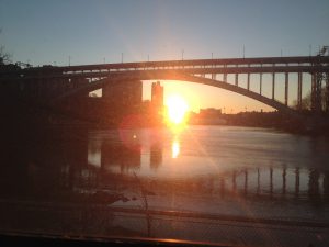 Photo of a sunrise over a body of water. A bridge span appears to be over the top of the sunrise.