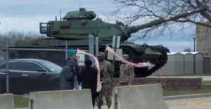 US Army tank in the background with military personnel and people with banners in front 