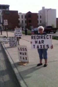 people with protest signs including “redirect war taxes,” “less war tax,” and “taxes for peace not war”
