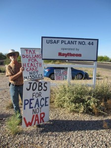 man holding signs reading “tax dollars for health care not missiles” and “jobs for peace not war” in front of a sign reading “U.S.A.F. plant #44, operated by Raytheon”